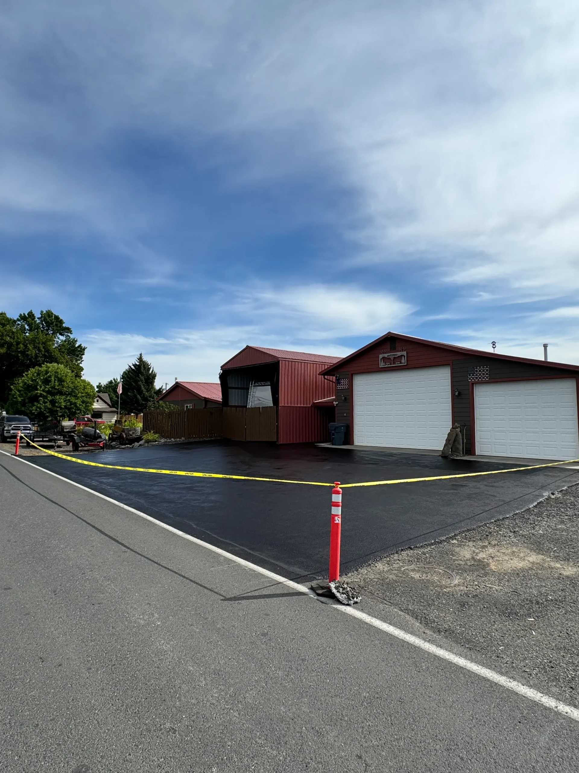 A freshly sealed black asphalt driveway in front of red residential garages, cordoned off with yellow caution tape