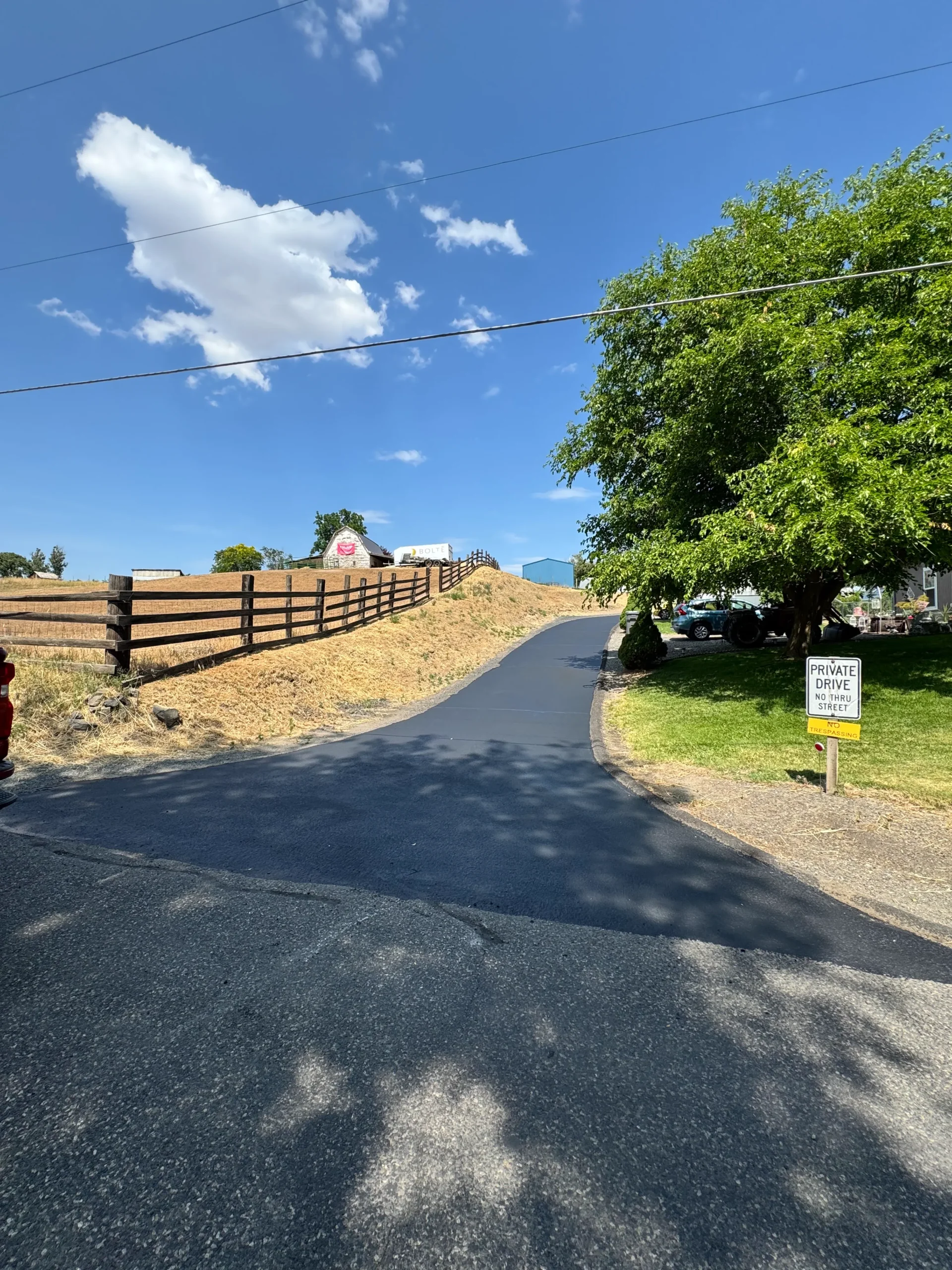 A newly sealed black asphalt driveway winding uphill toward a white barn with a Private Drive sign in the foreground