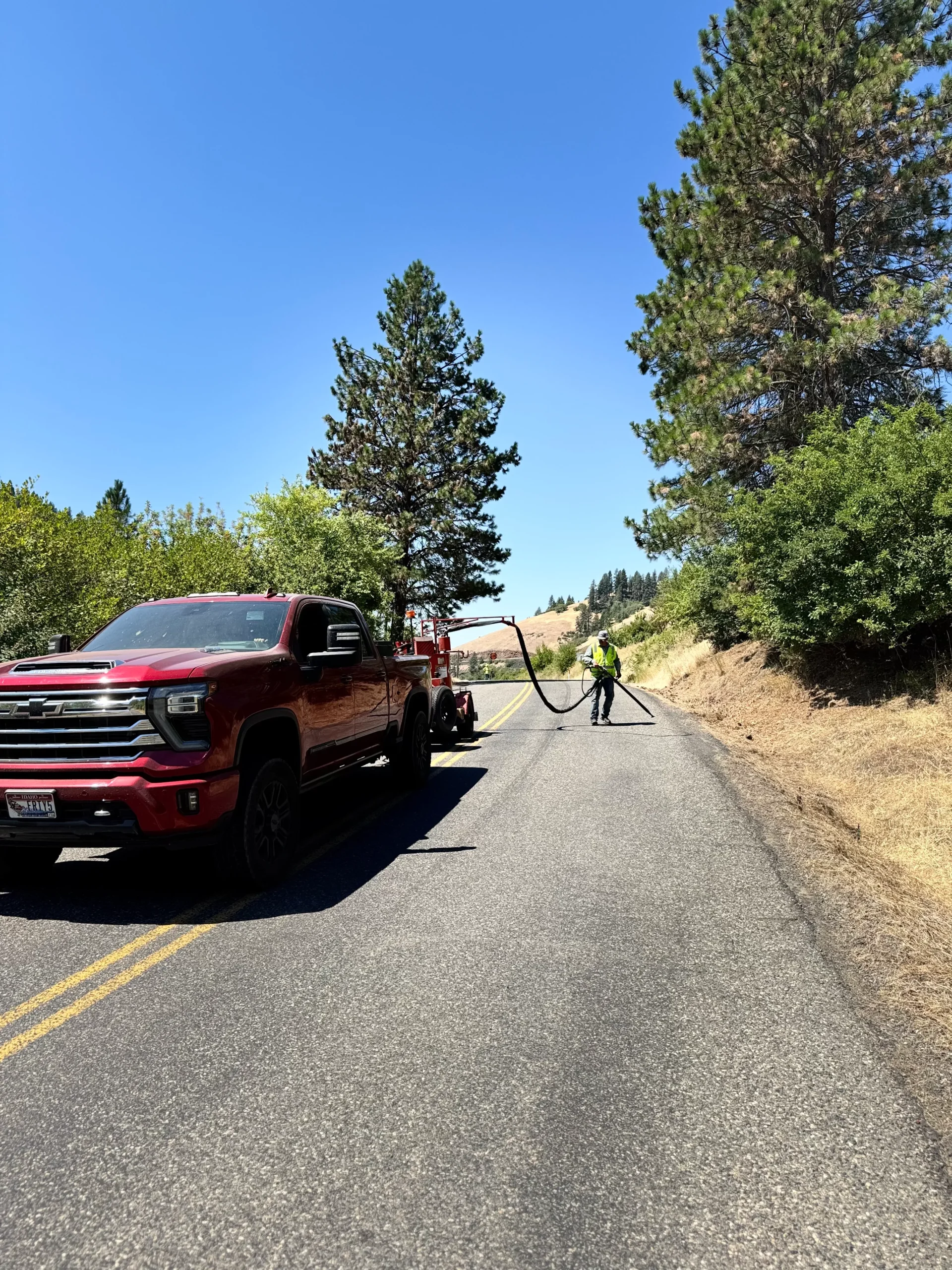 A red pickup truck parked on the side of a rural road as a worker uses a long hose to seal cracks in the pavement