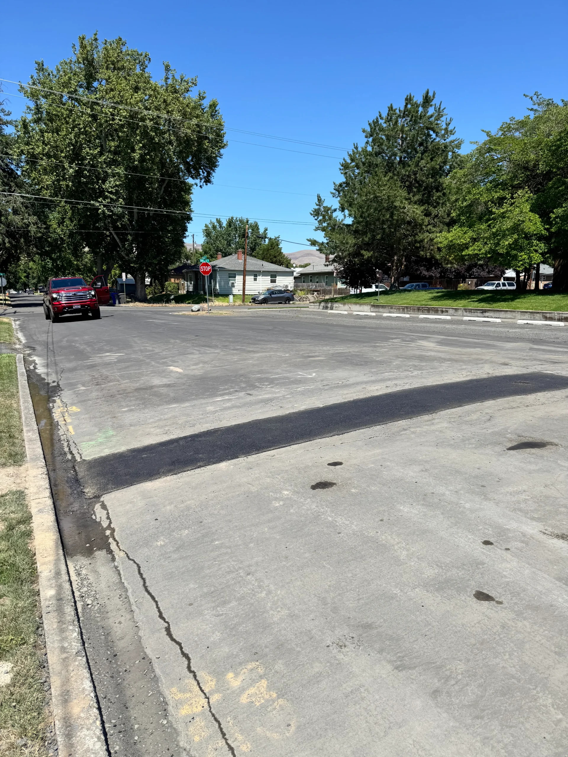 A wide asphalt road showing a fresh black patch across the lane with a red pickup truck parked in the background